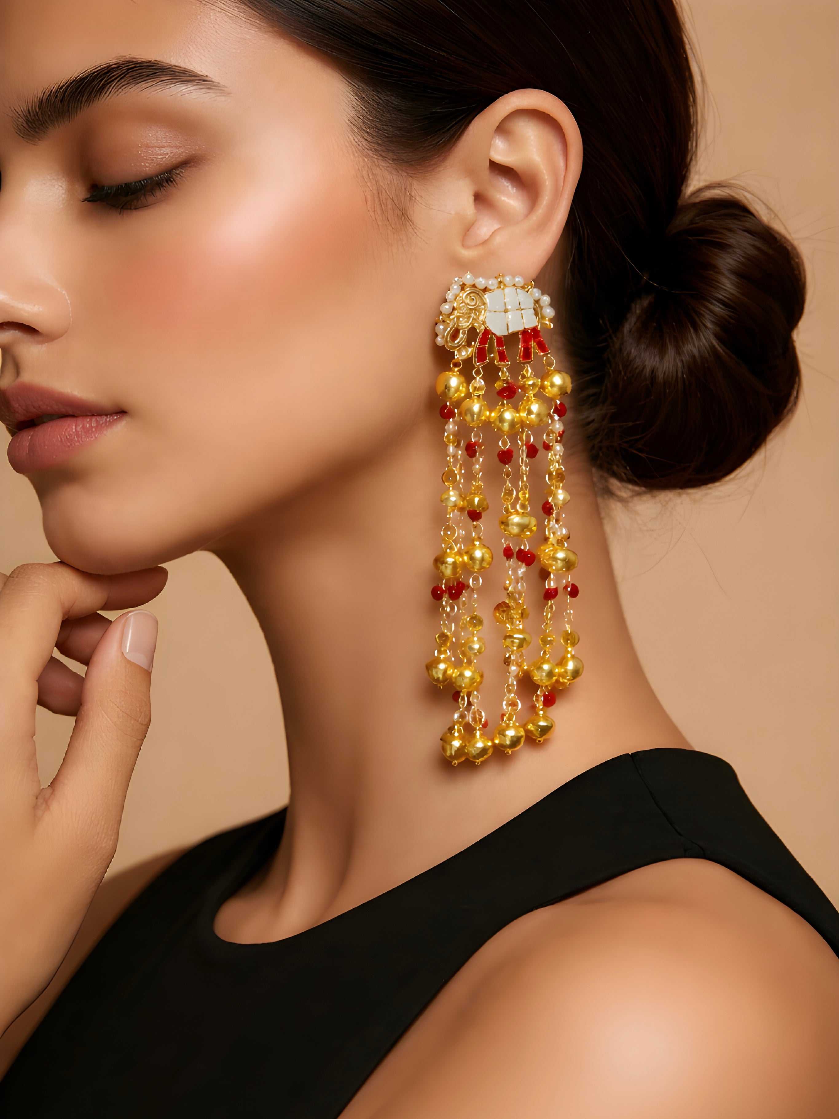 Close-up of a woman wearing a pair of long, dangling earrings with gold and red stones on a beige background.